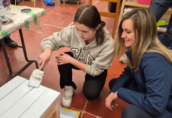 A teen camper painting a wooden bench while her aunt sits next to her