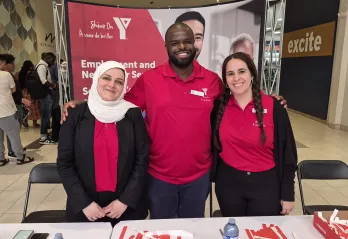 Three YMCA staff members standing and smiling together behind a booth promoting YMCA Employment and Newcomer Services at Merivale Mall.