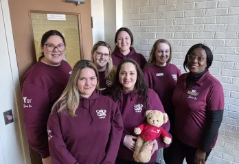 Group photo of seven YMCA Child Care team members wearing burgundy hoodies. One team member is holding a teddy bear wearing a Y Shine On shirt.