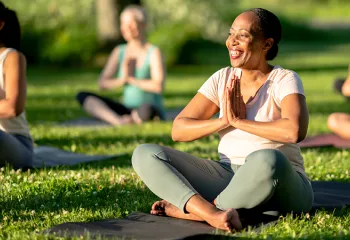 A woman smiles while sitting cross-legged on a yoga mat outdoors, hands in prayer position.