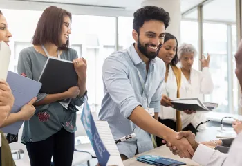 A diverse group of people in a job fair setting. A man in the foreground is smiling, shaking hands, and surrounded by others holding folders.