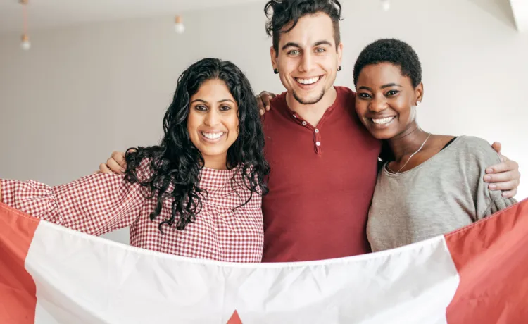 A man and two women, all of different cultural backgrounds, smiling and holding up a Canadian flag together