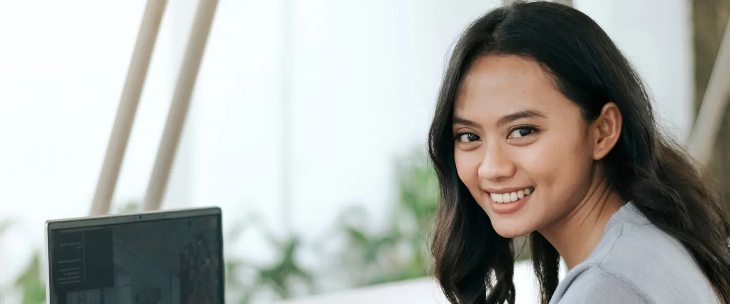 A young Asian woman smiles while working on her laptop.