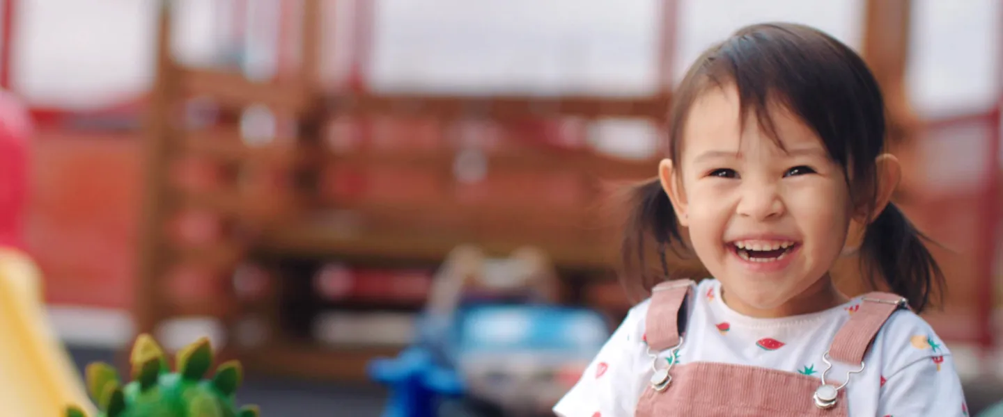 A smiling toddler sits at a table playing with toys.