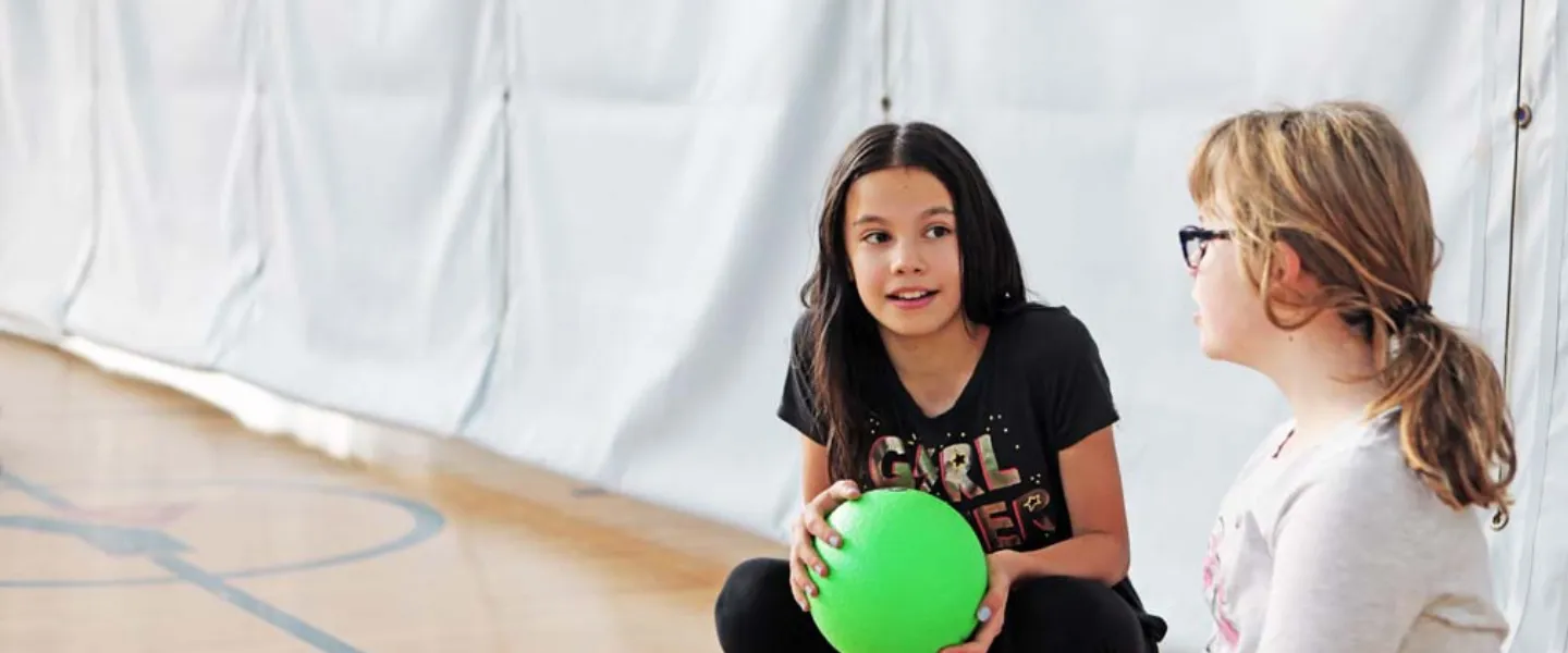 Two young girls sit in a gymnasium with a  ball.