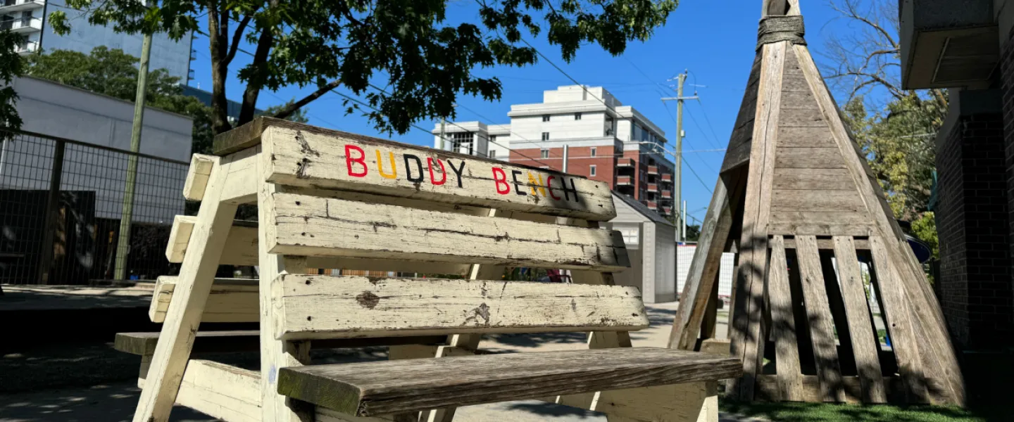 An outdoor play area. A wooden bench with the words "Buddy Bench" painted on it. A wooden tipi structure.