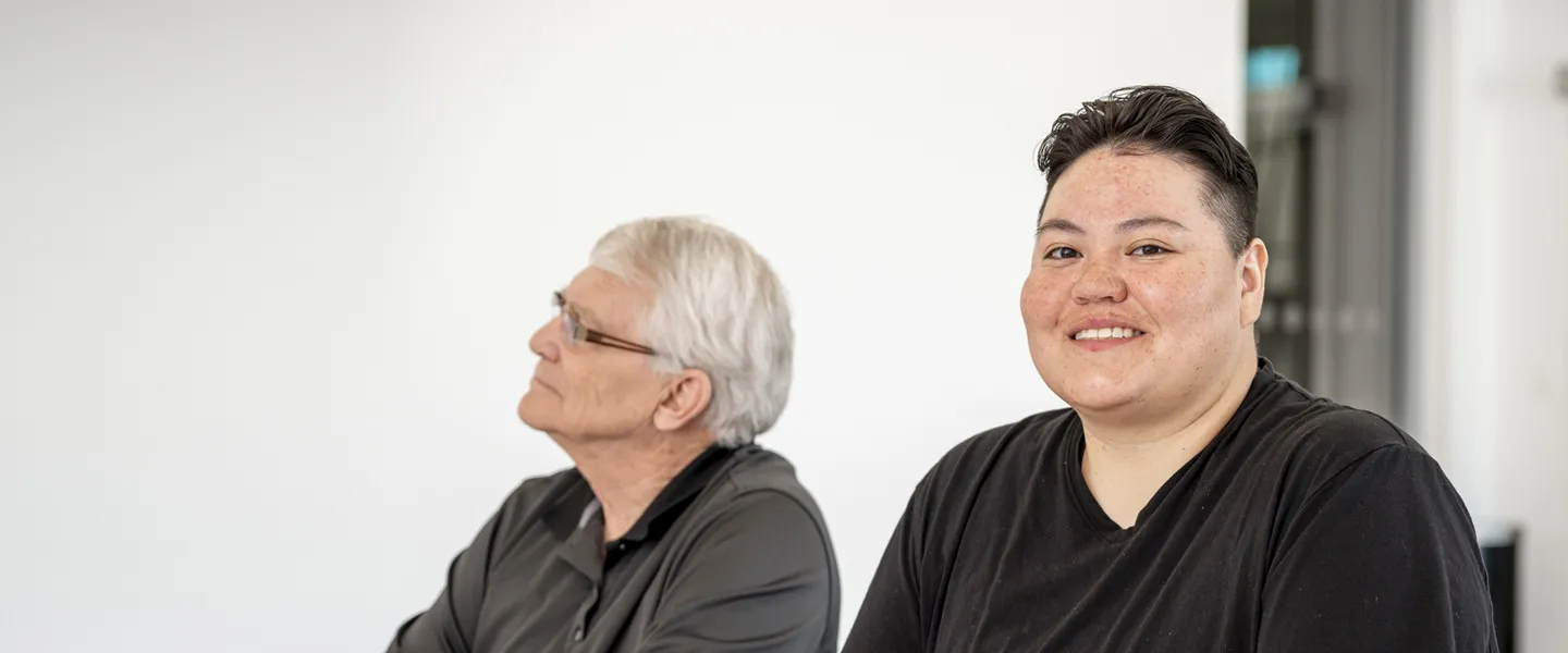 Two people sitting at a conference table. One person looks towards the presenter while the other one smiles at the camera.