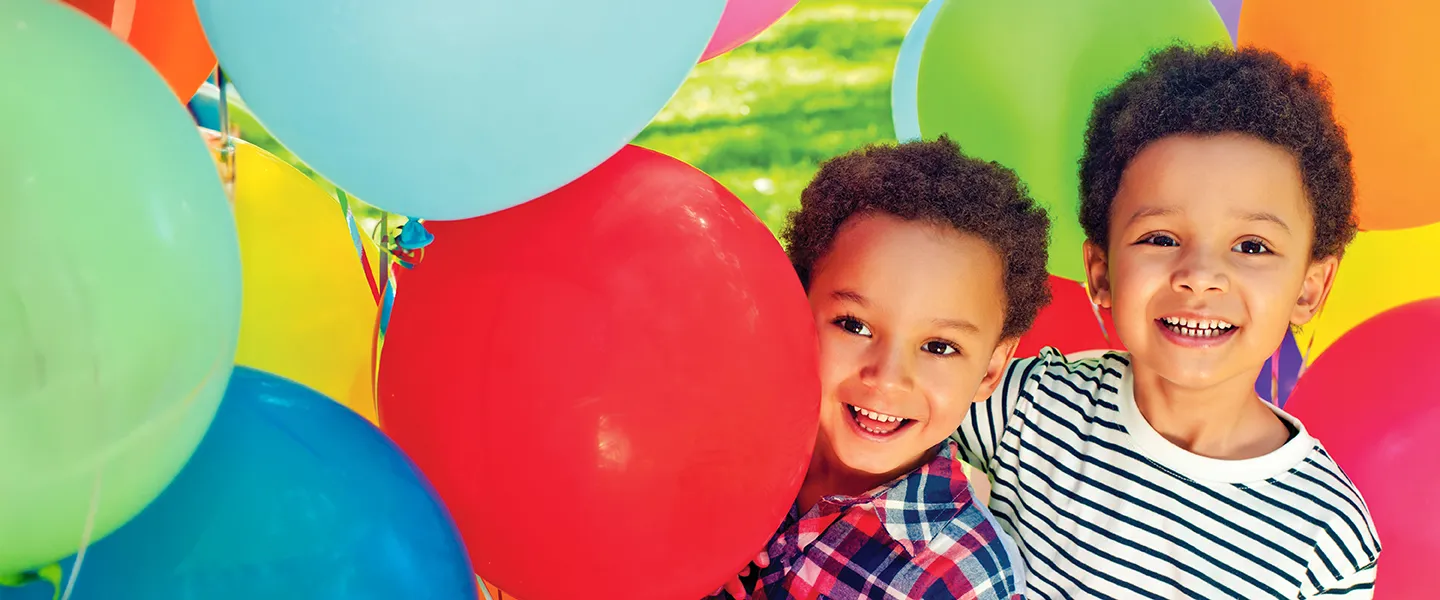 Two young boys smile joyfully in front of a vibrant display of colorful balloons.