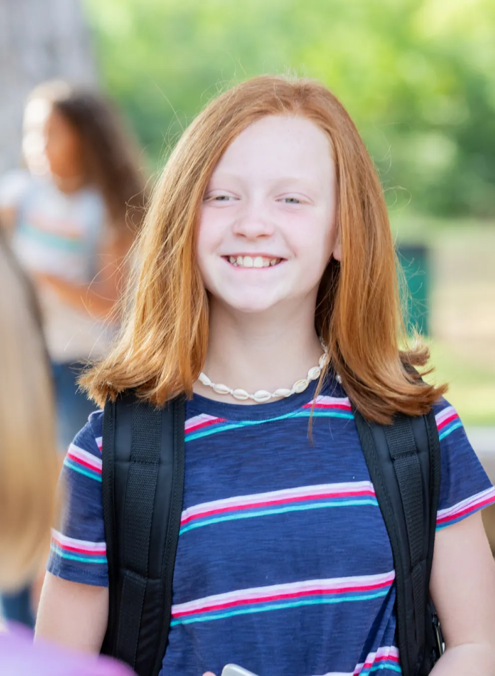 Two girls smiling at each other, wearing backpacks in a bright, outdoor setting