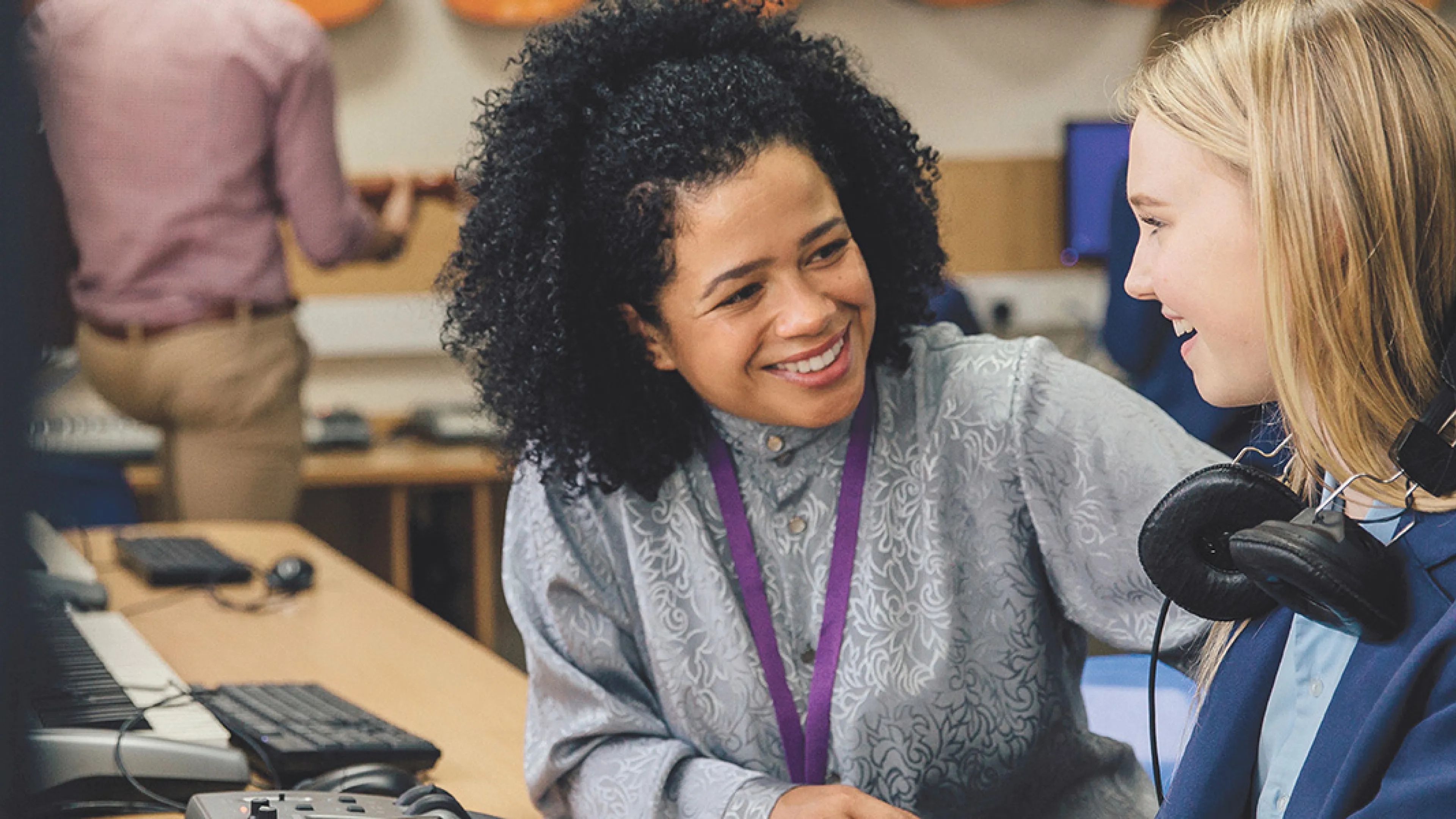 A young girl wearing headphones engages in conversation with her teacher in a classroom setting.
