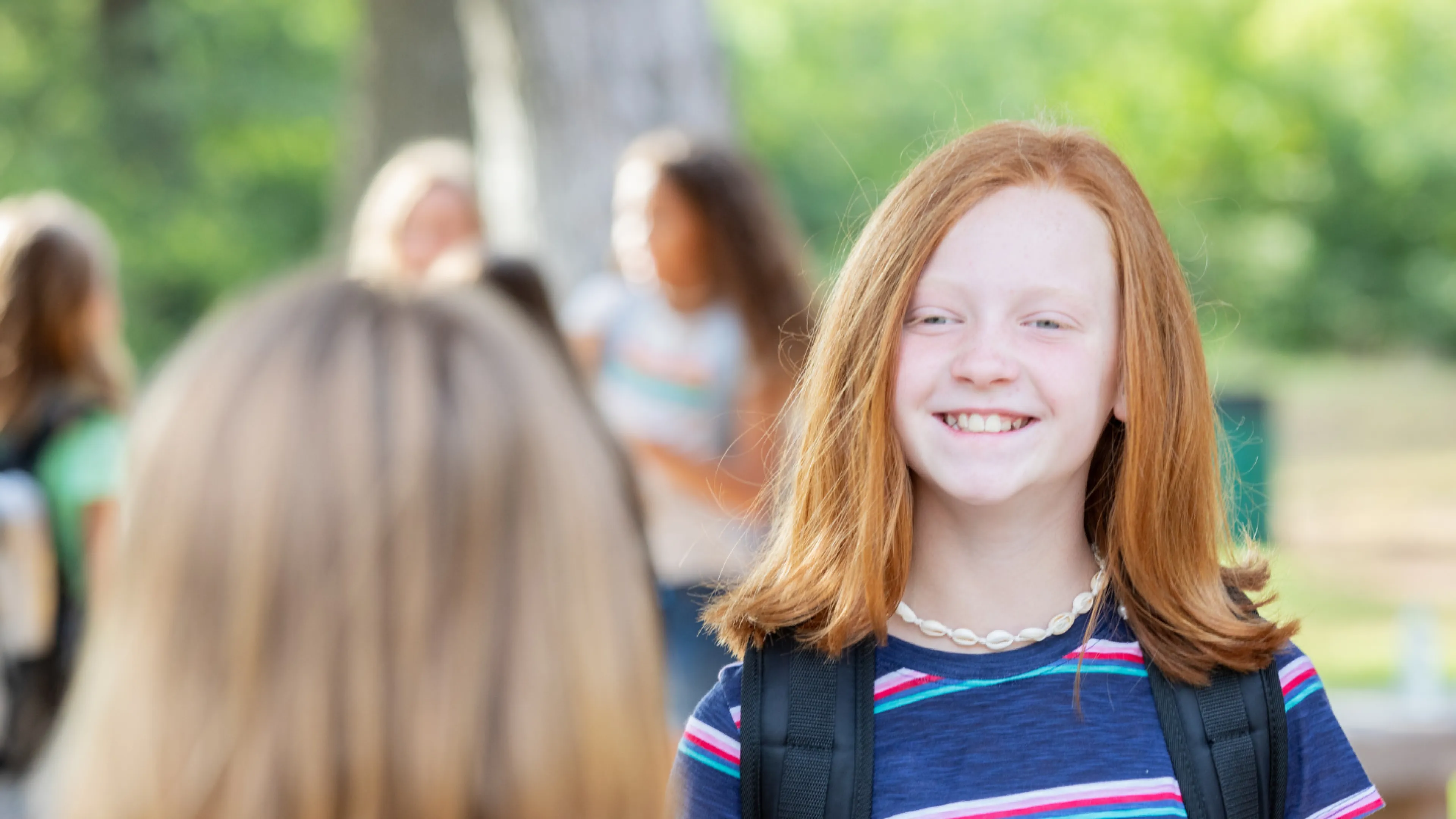 Two girls smiling at each other, wearing backpacks in a bright, outdoor setting
