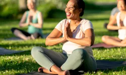 A woman smiles while sitting cross-legged on a yoga mat outdoors, hands in prayer position.