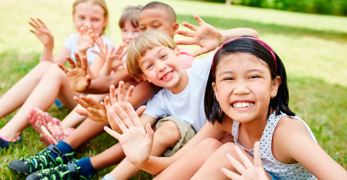 A group of children sit in a row smiling and waving.