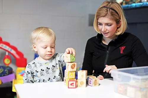 An early childhood educator in a Y logo jacket sitting with a child at a table. The child is stacking wooden blocks.