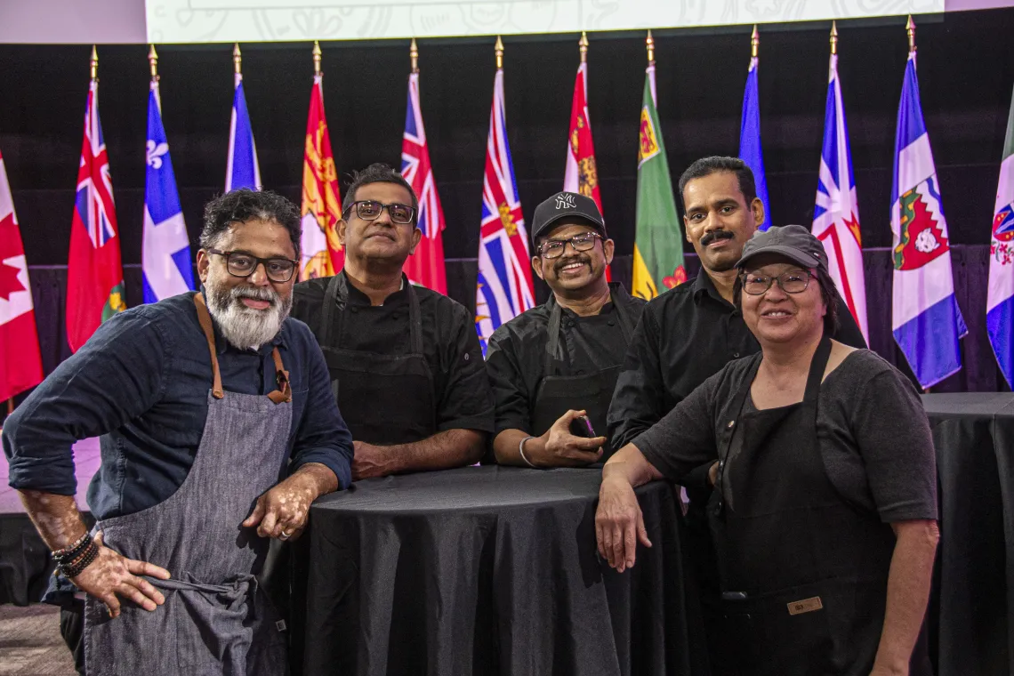 Five chefs leaning on a round table. There are a variety of flags in the background.