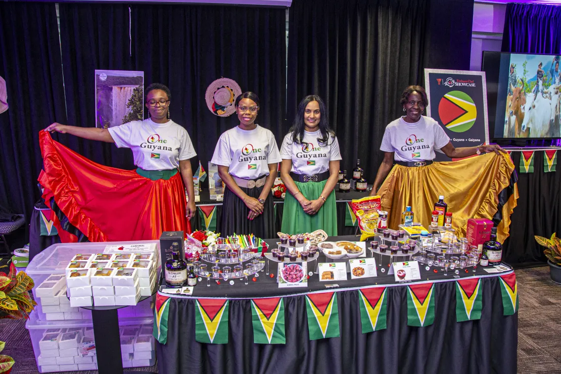 Guyana table covered in food and decorated with flags. Four women are standing behind the table, their skirts the colours of the Guyana flag.