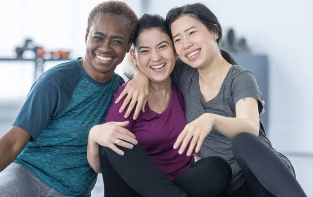 A group of three friends, including two women and one man, seated on the gym floor sharing a moment of camaraderie
