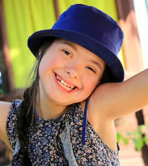 A girl wearing a blue hat, smiling and looking towards the camera in a sunny outdoor setting.
