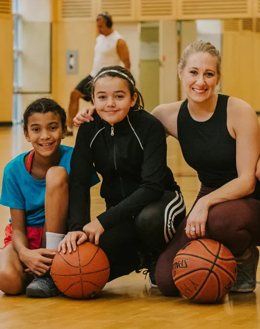 A diverse group of people sit smiling on a basketball court, each with a basketball.