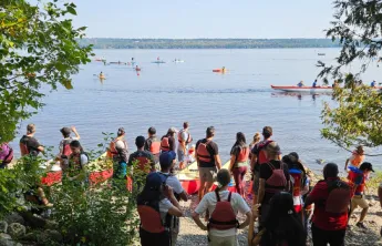 A group of people with red life jackets on. They are standing by red canoes on the Ottawa River shore and watching other people in canoes on the water.