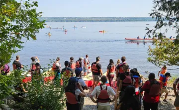 A group of people with red life jackets on. They are standing by red canoes on the Ottawa River shore and watching other people in canoes on the water.