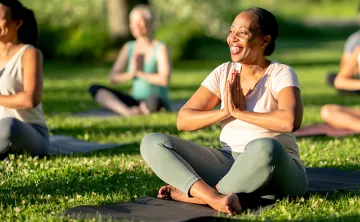 A woman smiles while sitting cross-legged on a yoga mat outdoors, hands in prayer position.