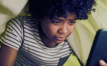 A child with curly hair, wearing a striped shirt, is intently looking at a smartphone under a blanket.