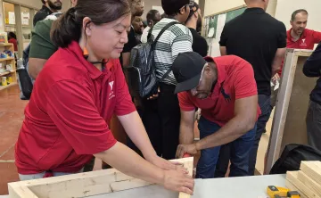 A woman holding wooden planks steady on a table as a man drills them together. There are several people in the background exploring skilled trades stations.