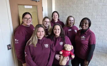 Group photo of seven YMCA Child Care team members wearing burgundy hoodies. One team member is holding a teddy bear wearing a Y Shine On shirt.