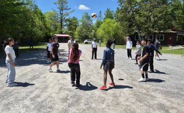 A group of newcomer youth playing with a volleyball on gravel. They are surrounded by a forested area.